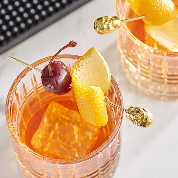 Two glasses of orange juice with Barfly gold cocktail picks holding cherries and orange slices.
