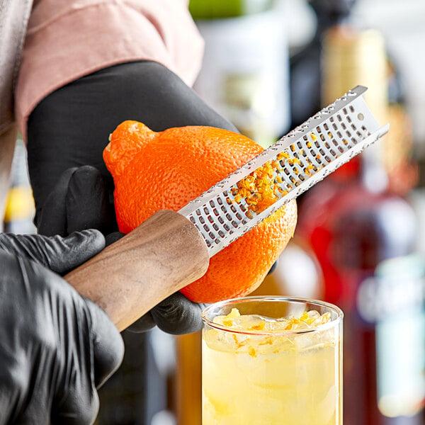 A person in black gloves using a Barfly zester grater to peel an orange over a drink.