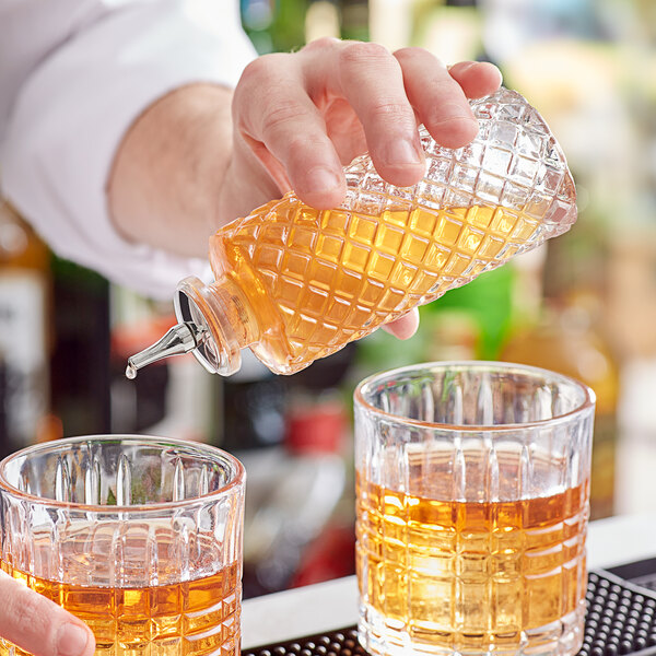 A hand pouring Barfly glass bitters bottles into glasses of amber liquid.