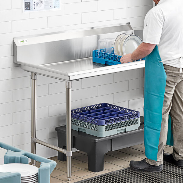 A man in a blue apron standing next to a Regency stainless steel clean dish table with a sink.