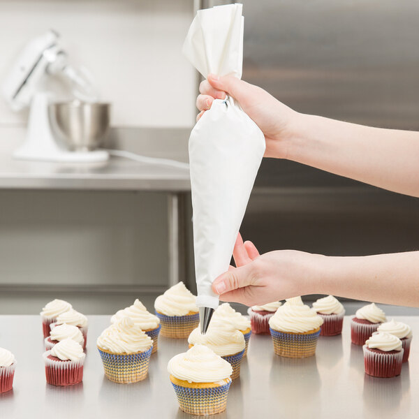 A person's hand holding a white Ateco pastry bag over a cupcake with white frosting.