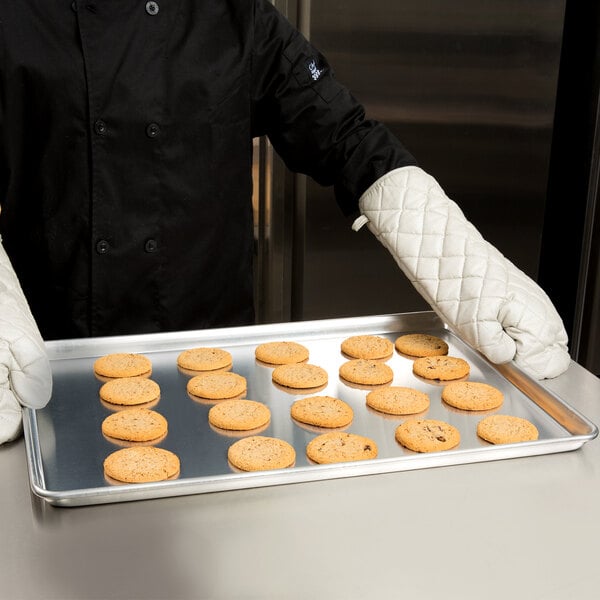 A person wearing a chef's uniform and gloves holding a Vollrath Wire in Rim Aluminum Bun Pan full of cookies.