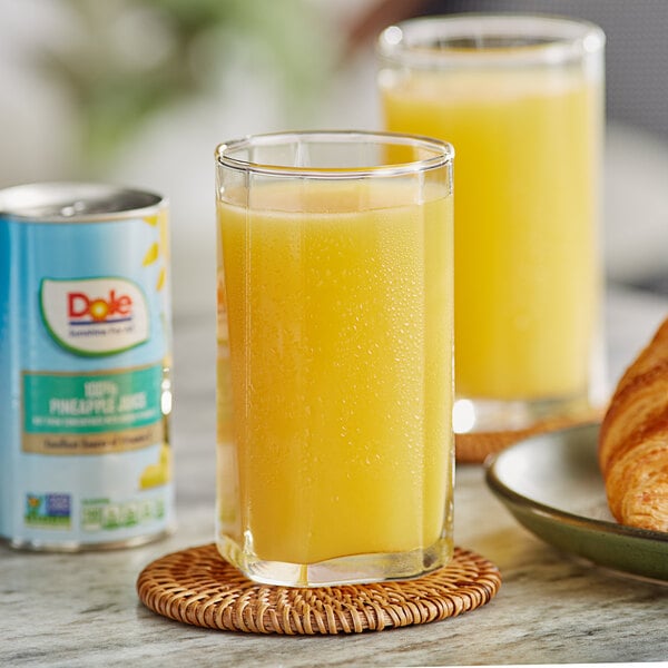 A glass of Dole pineapple juice next to a croissant on a table.