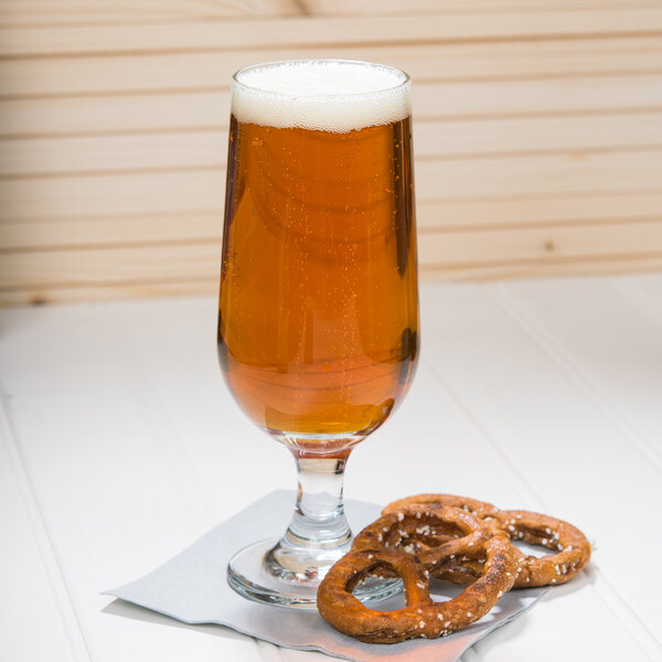 A Libbey stemmed pilsner glass of beer and pretzels on a white table.