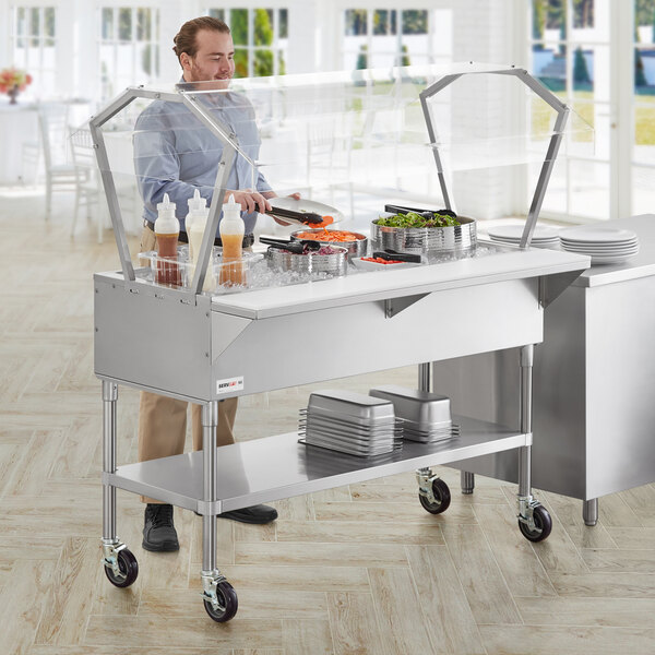 A man standing behind a ServIt stainless steel ice-cooled food table with a sneeze guard.