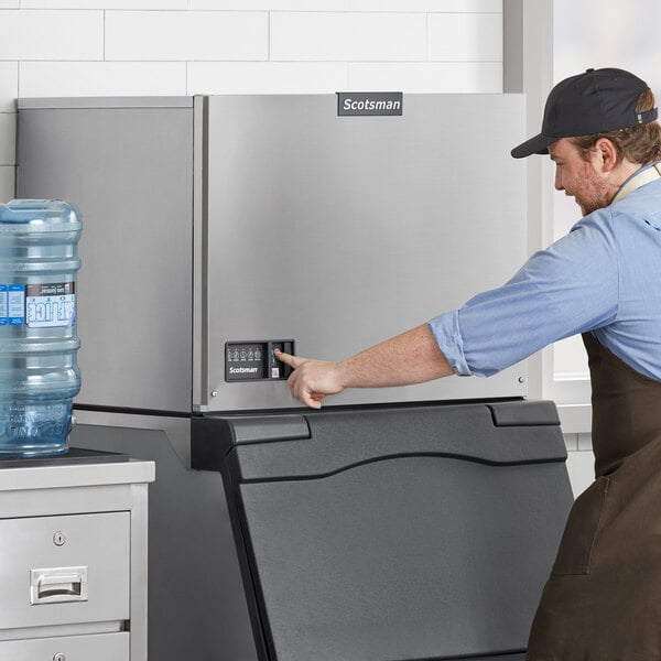 A man in a brown apron and cap pressing a button on a Scotsman water cooled ice machine.