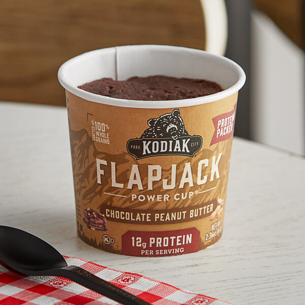 A Kodiak Cakes chocolate peanut butter flapjack cup on a table with a spoon and red and white checkered cloth.