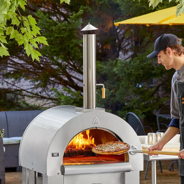 A man using a Backyard Pro stainless steel wood-fired pizza oven to cook a pizza.