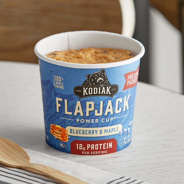 A blue container of Kodiak Cakes Blueberry and Maple Flapjack Cup mix with a spoon inside on a table.