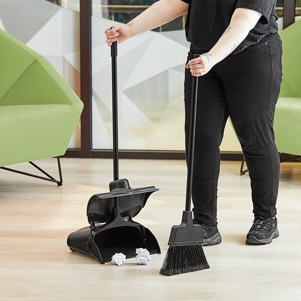 A woman holding a Lavex closed-lid lobby dust pan with broom sweeping the floor.