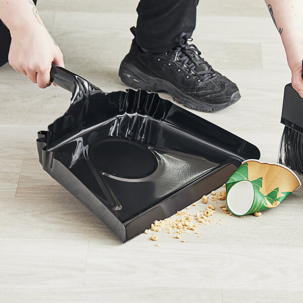 A person using a broom and Lavex black steel dust pan to sweep a floor.