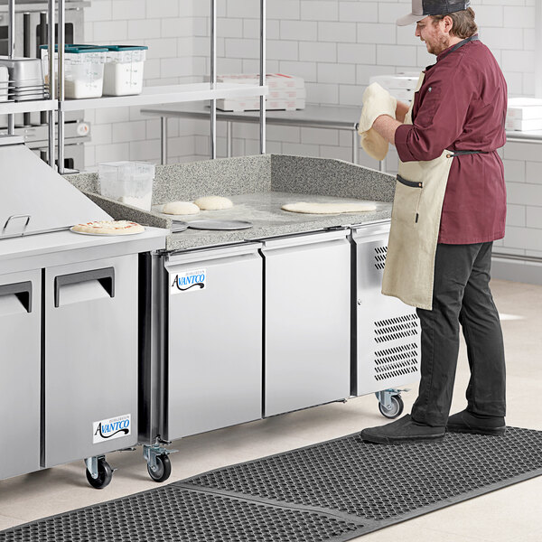 An Avantco 2 door stone top refrigerated pizza prep table on a counter in a professional kitchen with a person wearing an apron.