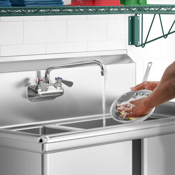 A person washing dishes with a Regency wall mount faucet over a stainless steel sink.