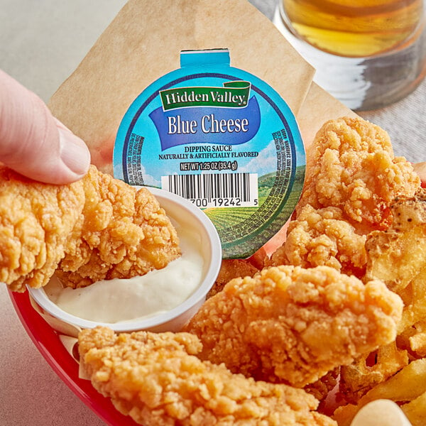 A hand holding a bowl of Hidden Valley Blue Cheese dressing over a plate of fried chicken.