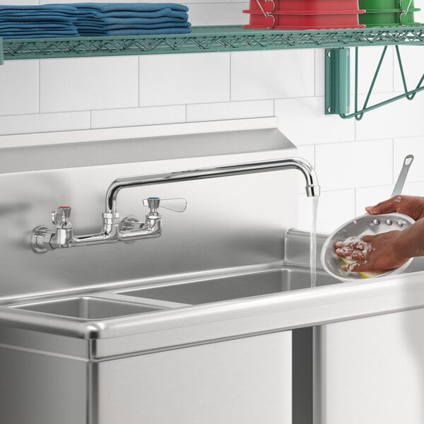 A person using a Regency wall mount faucet to wash dishes in a stainless steel sink.