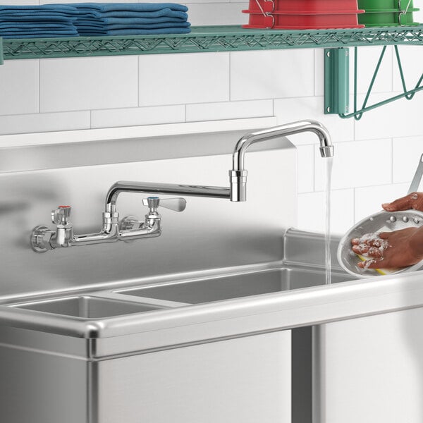 A person using a Regency wall mount faucet with double-jointed spout to wash dishes in a stainless steel sink.