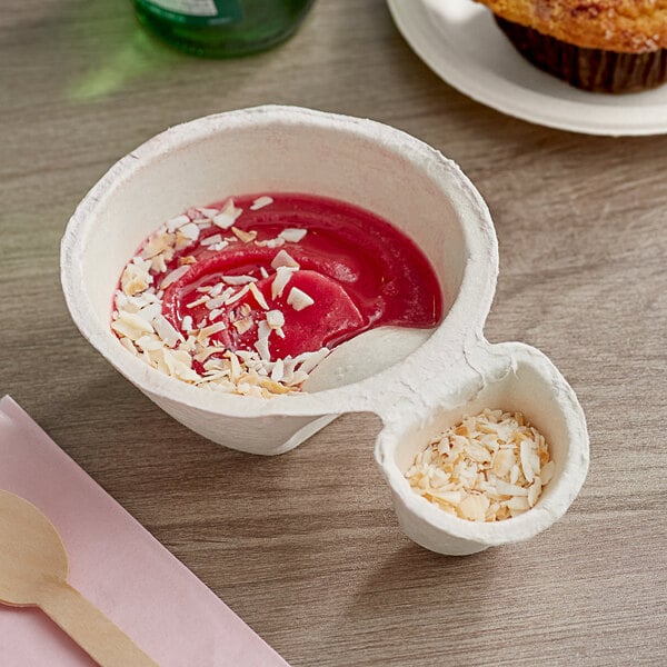 A white bowl of red liquid with coconut shavings on a table with a wooden spoon.