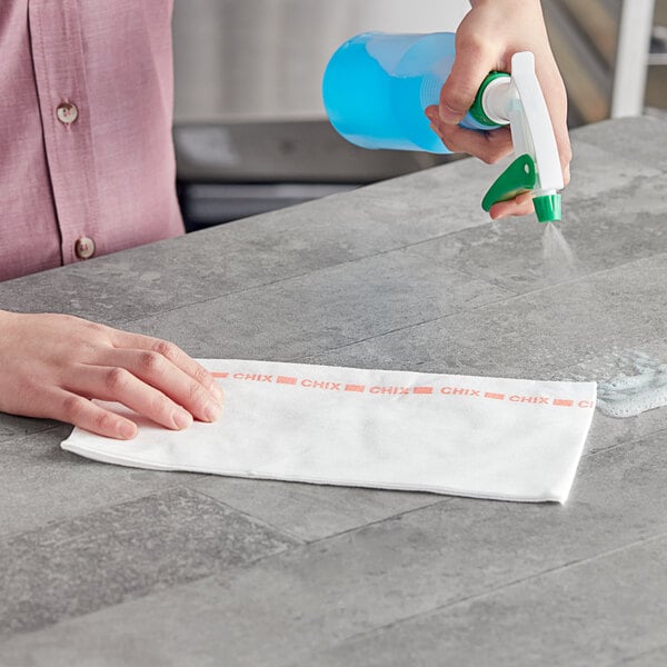A person using a blue spray bottle to clean a counter with a white Chix foodservice towel.
