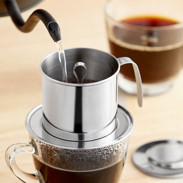 A stainless steel Vietnamese coffee press pouring coffee into a metal cup.