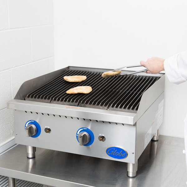 A person in a white coat cooking food on a Globe gas charbroiler with chicken on it.
