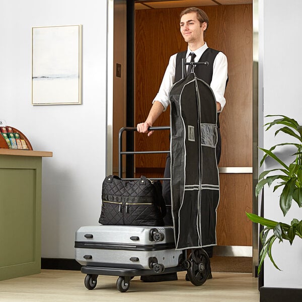A man pushing a Lancaster Table & Seating black luggage cart with a black bag on top.