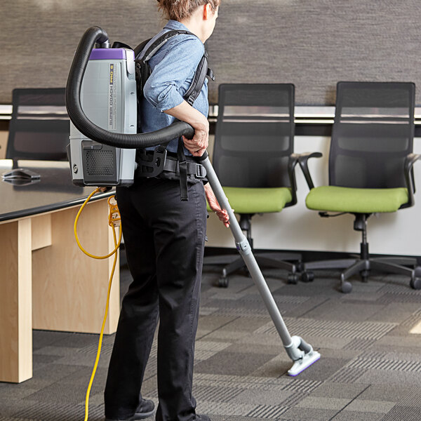 A woman wearing a backpack vacuuming a green office chair with armrests.