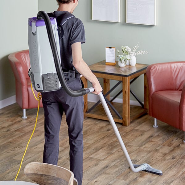 A man in black and grey using a ProTeam backpack vacuum to clean a wood floor.