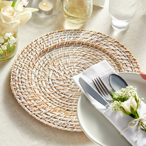 A white plate with a white napkin and Acopa blond rattan charger plate with silverware on it.