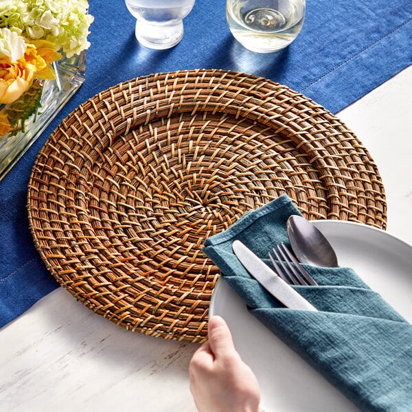 A hand placing a spoon and fork on a woven Acopa rattan charger plate.