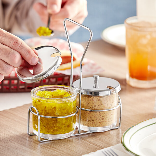A person holding a spoon with mustard pouring it into a glass jar in a wire condiment caddy.