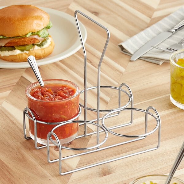 A metal wire condiment holder with a jar of sauce next to a burger.