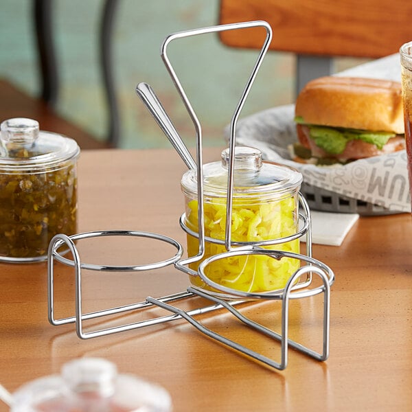 A metal wire condiment holder on a table with a sandwich and jars of mustard and pickles inside.