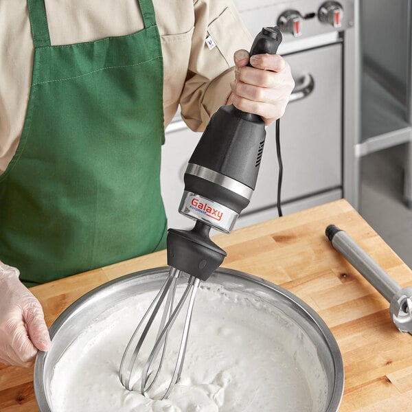 A person in a green apron using a Galaxy immersion blender with a whisk attachment to mix white liquid in a bowl.