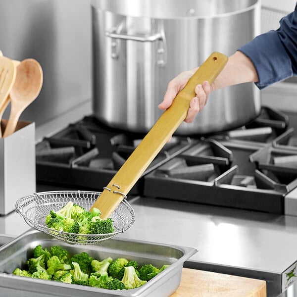 A person using an Emperor's Select fine skimmer with a bamboo handle to stir a bowl of broccoli.
