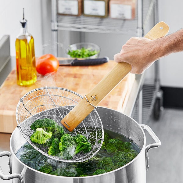 A person using an Emperor's Select wooden skimmer to stir broccoli in a pot.