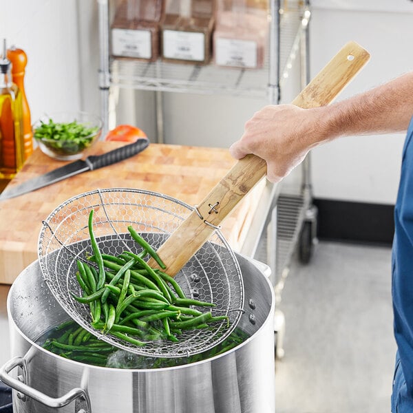 A person using Emperor's Select Coarse Skimmer with Bamboo Handle to stir green beans in a pot.