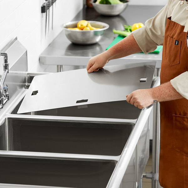 A person in an apron cutting a piece of metal for a Regency stainless steel sink cover on a counter in a professional kitchen.