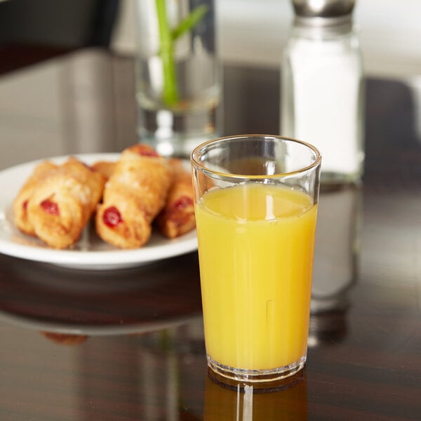 A Carlisle clear plastic tumbler filled with orange juice on a table next to a plate of pastries.