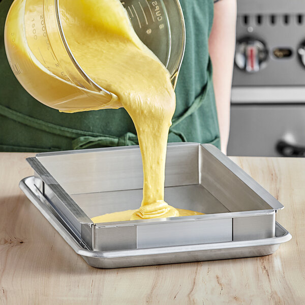 A person pouring yellow liquid into a Baker's Mark sheet pan using a metal device.