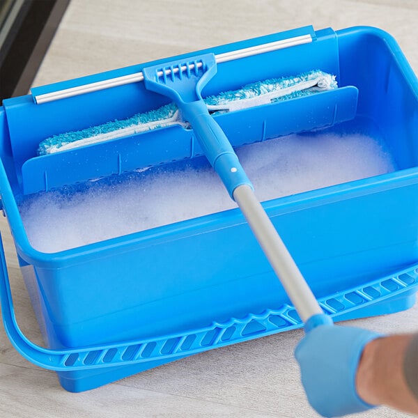 A hand using a mop to clean a window with a blue bucket filled with foam.