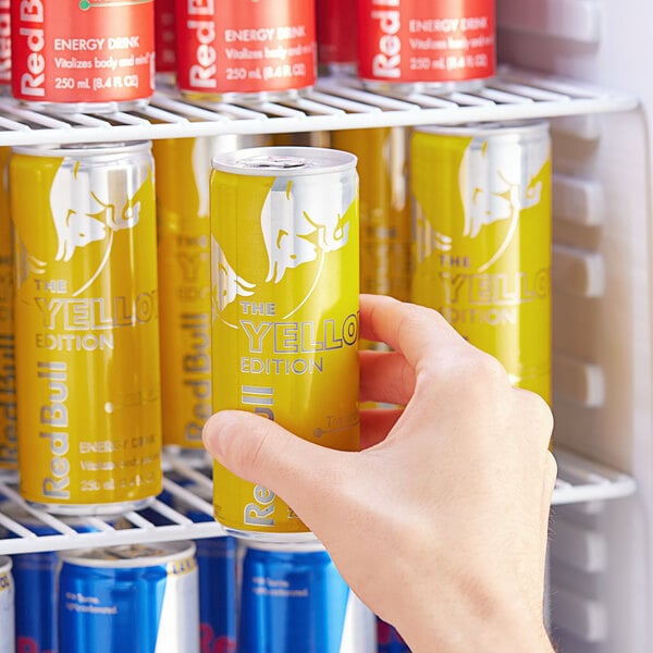 A hand taking a yellow and blue Red Bull Tropical energy drink out of a refrigerator.