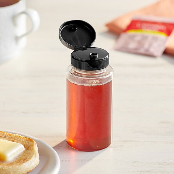 A 5.5 oz. clear cylinder sauce bottle with black cap on a table with breakfast items.