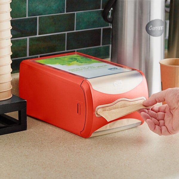 A hand taking a red napkin from a Tork Xpressnap countertop dispenser.