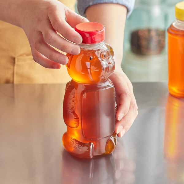 A person pouring honey into a bear-shaped plastic bottle on a kitchen counter.