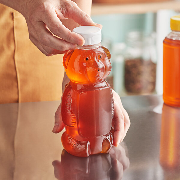 A hand using a white plastic dispensing cap to pour honey into a plastic bear bottle.