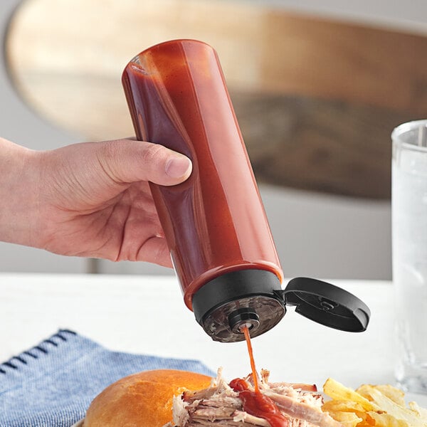 A hand holding a clear cylinder bottle of ketchup with a black lid pouring ketchup onto a sandwich.