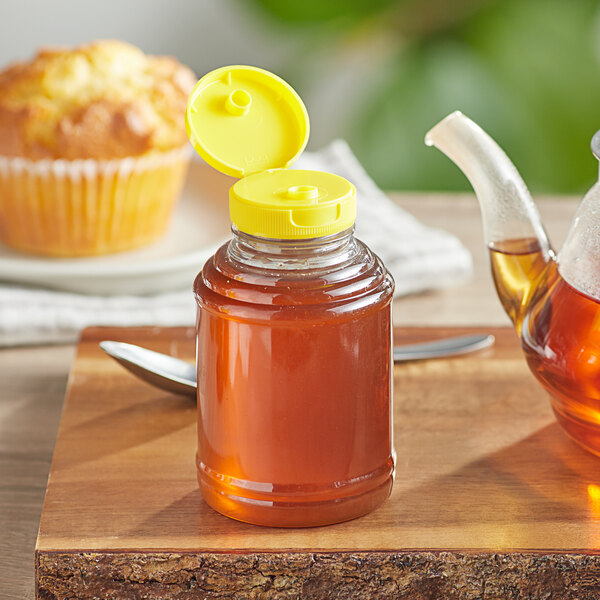 A close-up of a jar of honey with a yellow plastic flip top lid next to a muffin.