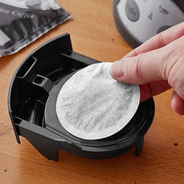 A hand holding a round white paper on a black plastic container with a picture of a coffee filter.