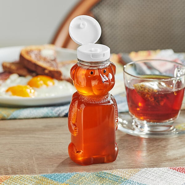A bear-shaped PET honey bottle with a white flip top lid on a table next to a cup of tea and toast.