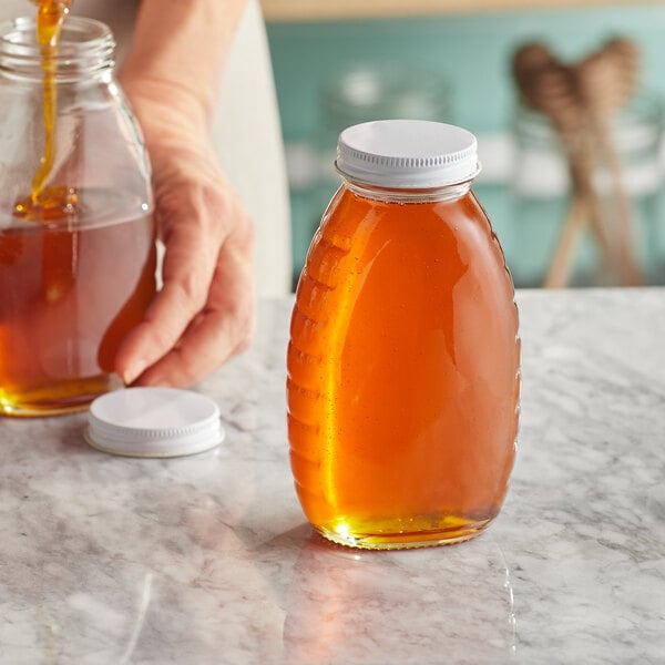 A person pouring honey into a classic glass honey jar with a white metal lid.